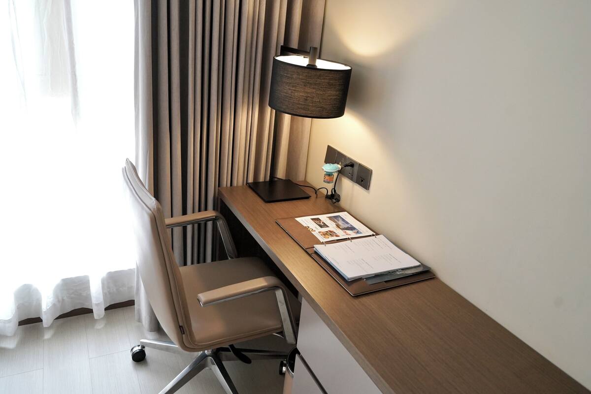 A hotel room with a wooden desk, a chair, a lamp, and a window with curtains.