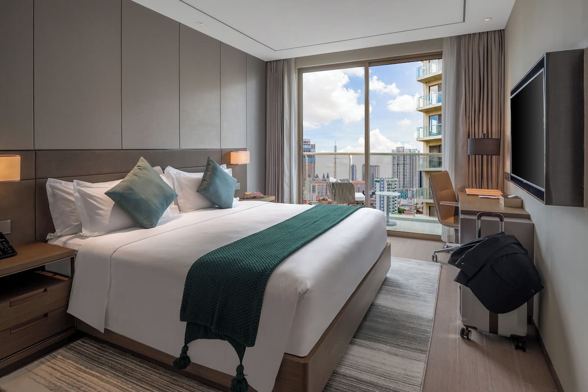 A modern hotel room with a large bed, a desk, a TV, and a view of the city skyline.