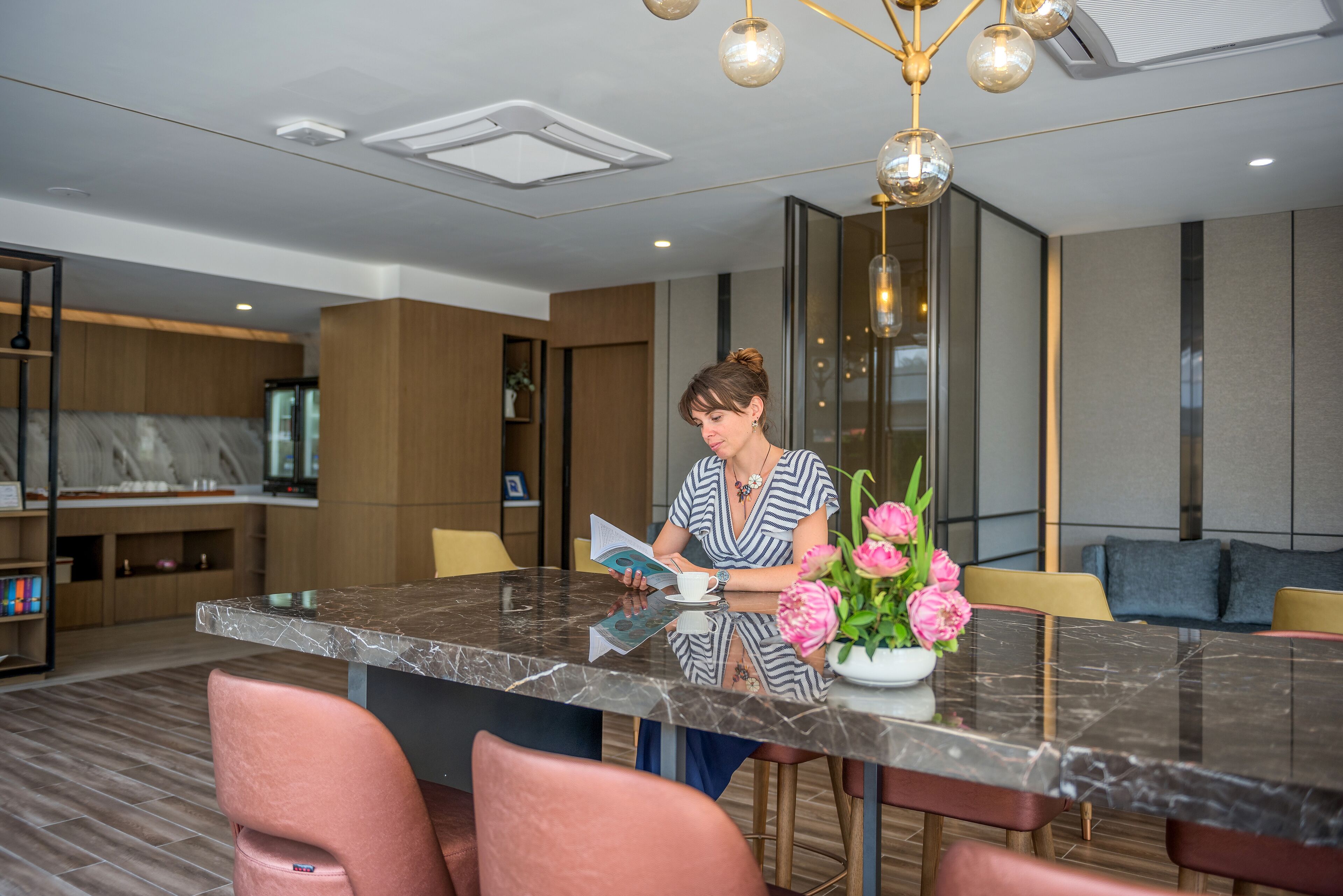 A modern interior with a marble dining table, pink chairs, and a woman reading a magazine.