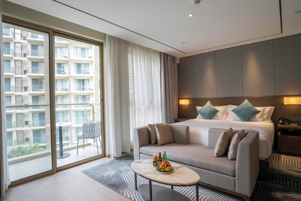 A modern hotel room with a large bed, a sofa, a small table with drinks, and a view of a high-rise building.