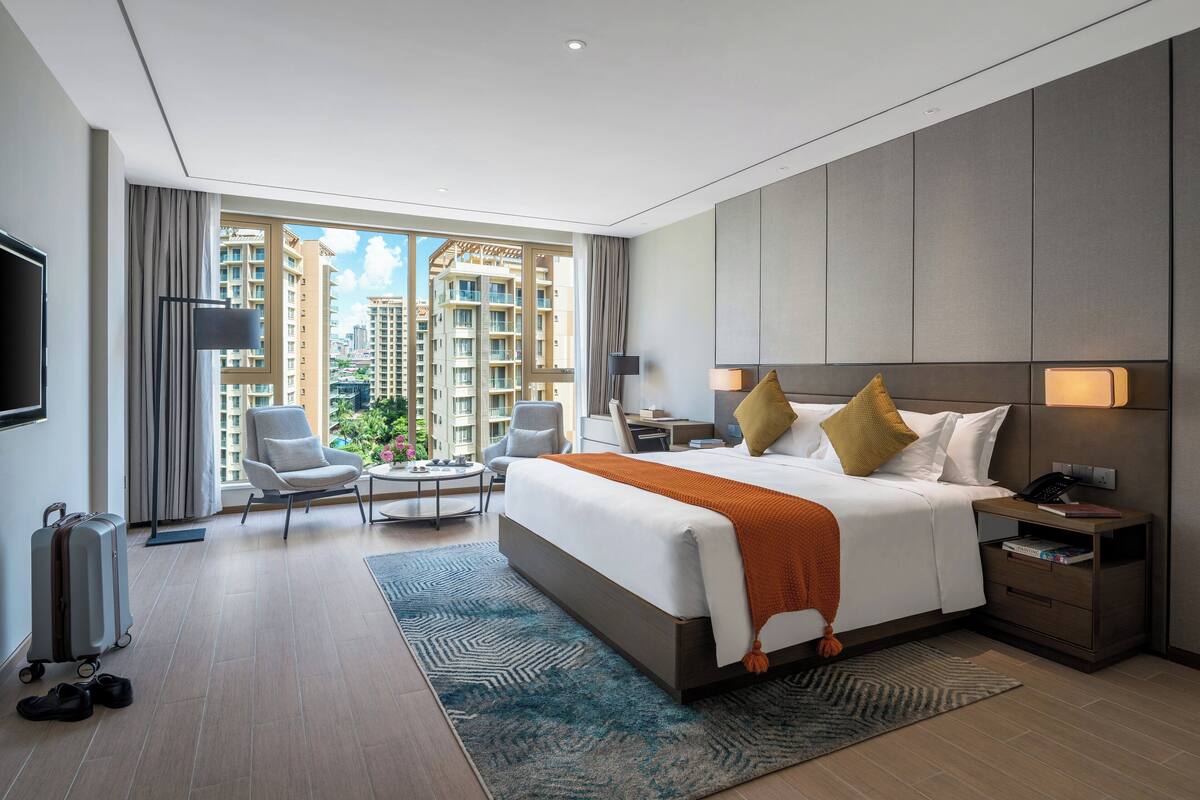 A modern hotel room with a large bed, a desk, a chair, and a view of high-rise buildings.