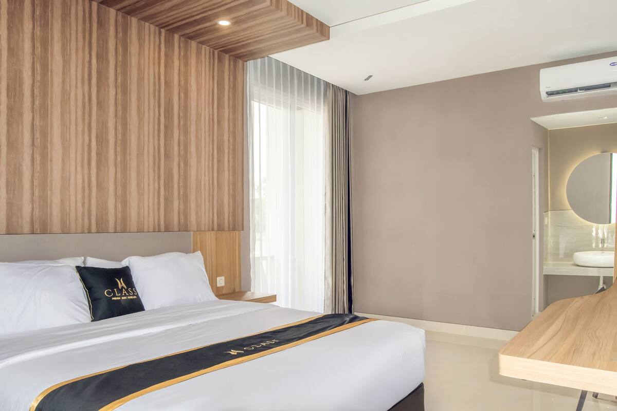 A hotel room with a large bed, a wooden headboard, a bedside table, a mirror, and a window with curtains.