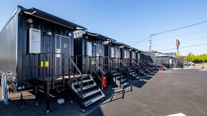 A row of modular, industrial-style buildings with external staircases, numbered rooms, and a parking area.