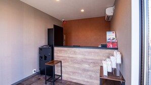 A hotel reception area with a wooden reception desk, a small table, a black filing cabinet, and a wall-mounted air conditioner.