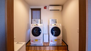 A laundry room with two stacked front-loading washing machines, a small window, and an air conditioning unit on the wall. - HOTEL R9 The Yard Isa (Isa)