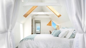Une chambre avec un plafond en pente, un lit recouvert de draps blancs et une vue sur la mer par une porte ouverte.