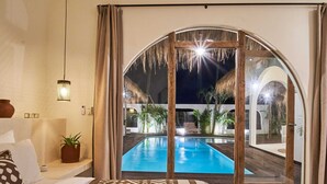 A bedroom with a large bed, a bedside table, a potted plant, and a view of an outdoor swimming pool at night. - Stunning 4BR Villa With Rice Field View in Umalas (Kerobokan)