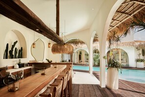 Uma área de bar com móveis de madeira, piscina e plantas tropicais.