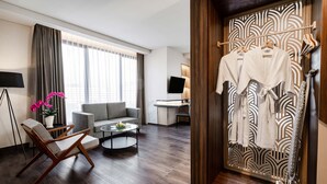 A modern hotel room with a sofa, a coffee table, a chair, a TV, and a wardrobe with robes. - Quest Prime Pemuda - Semarang (Semarang)