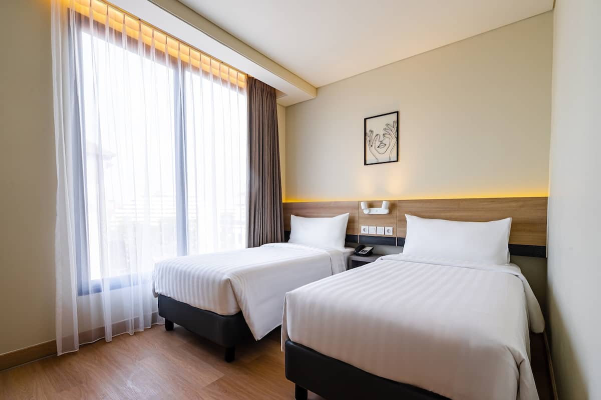 Habitación de hotel con dos camas, cabecero de madera, mesita de noche con teléfono y ventana con cortinas translúcidas.