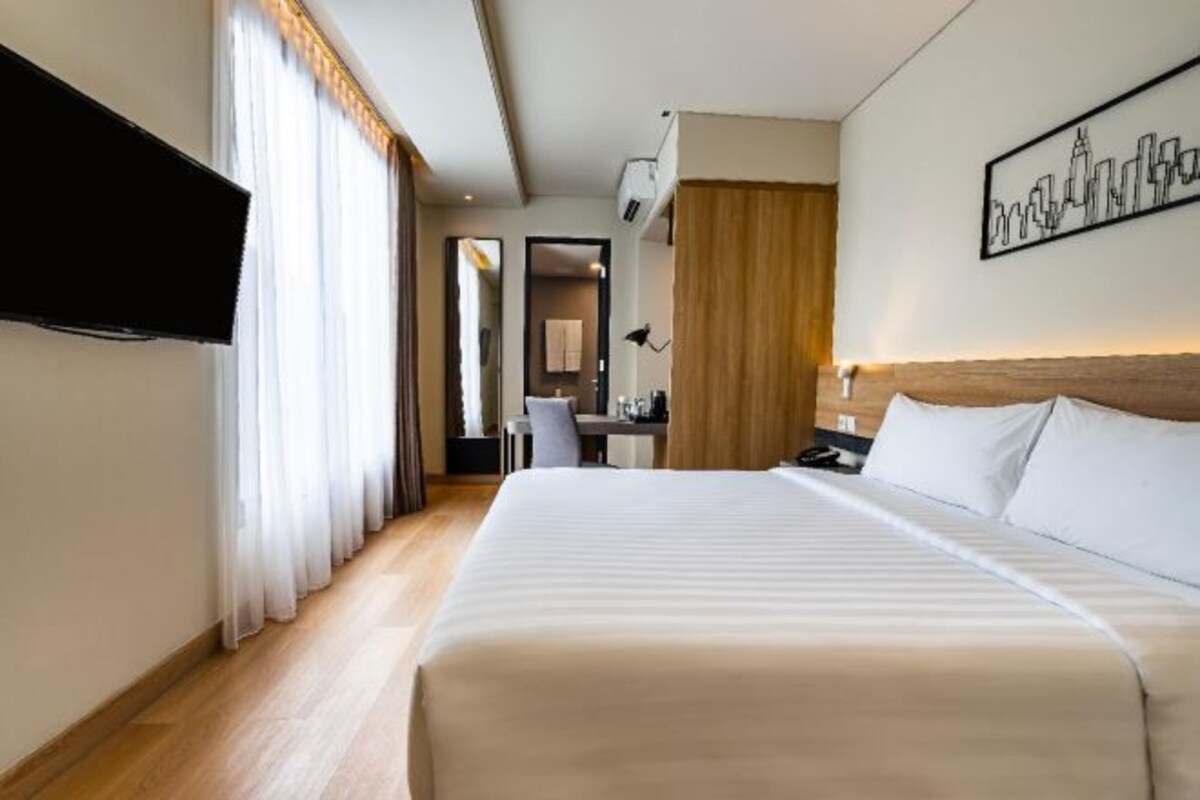 Quarto de hotel com uma cama grande, televisão de tela plana na parede, escrivaninha com cadeira e uma obra de arte com vista para a cidade na parede.