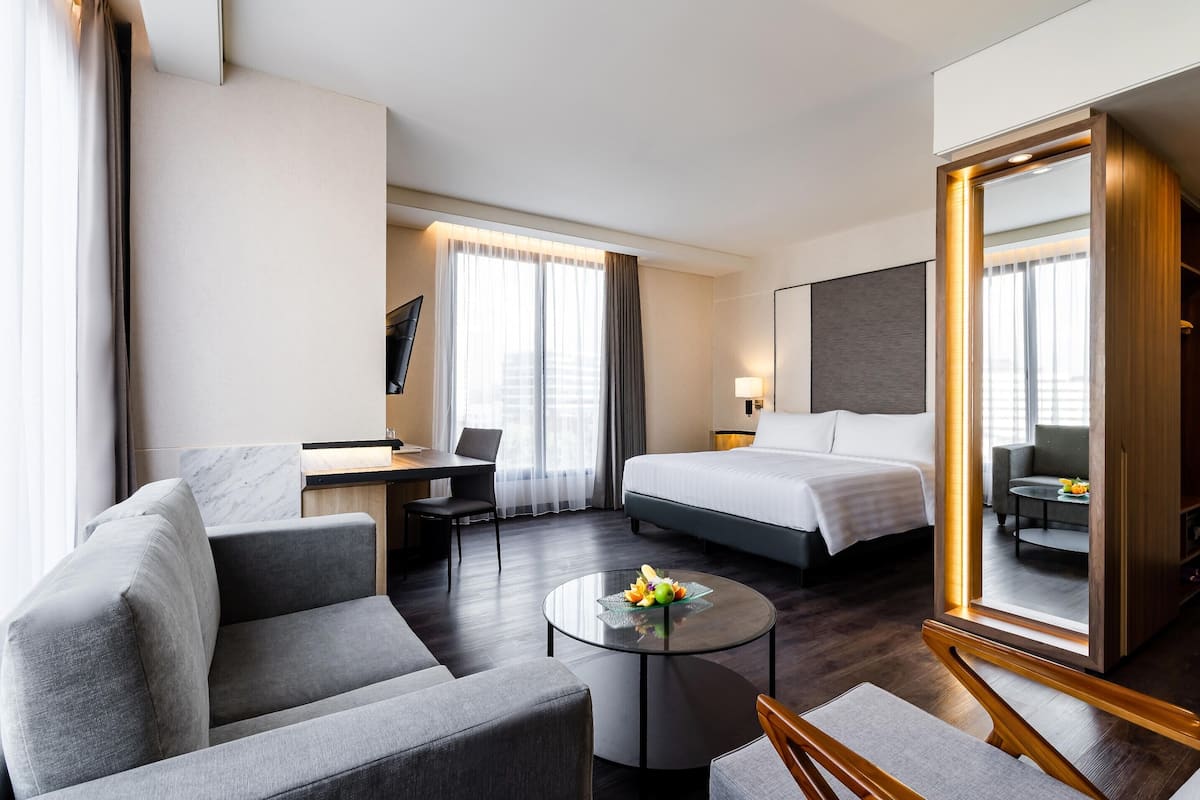 Una habitación de hotel con cama, sofá, escritorio, una mesa redonda de centro con fruta y un espejo grande.