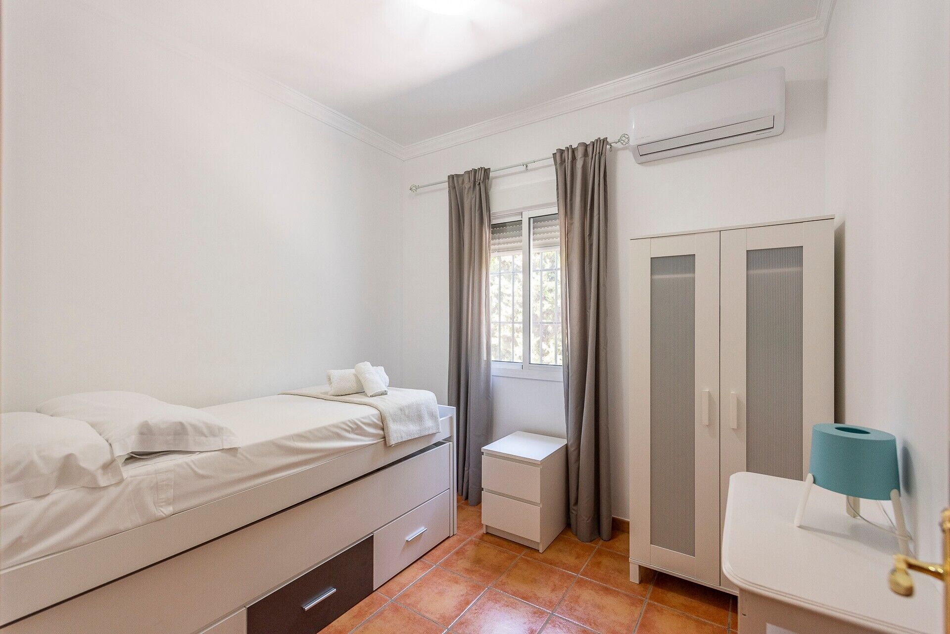 A bedroom with a bed, a nightstand, a dresser, a window with curtains, and a radiator.