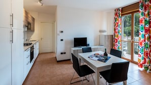 Una cocina moderna con armarios blancos, una zona de comedor con mesa y sillas, un televisor y una ventana con cortinas coloridas.