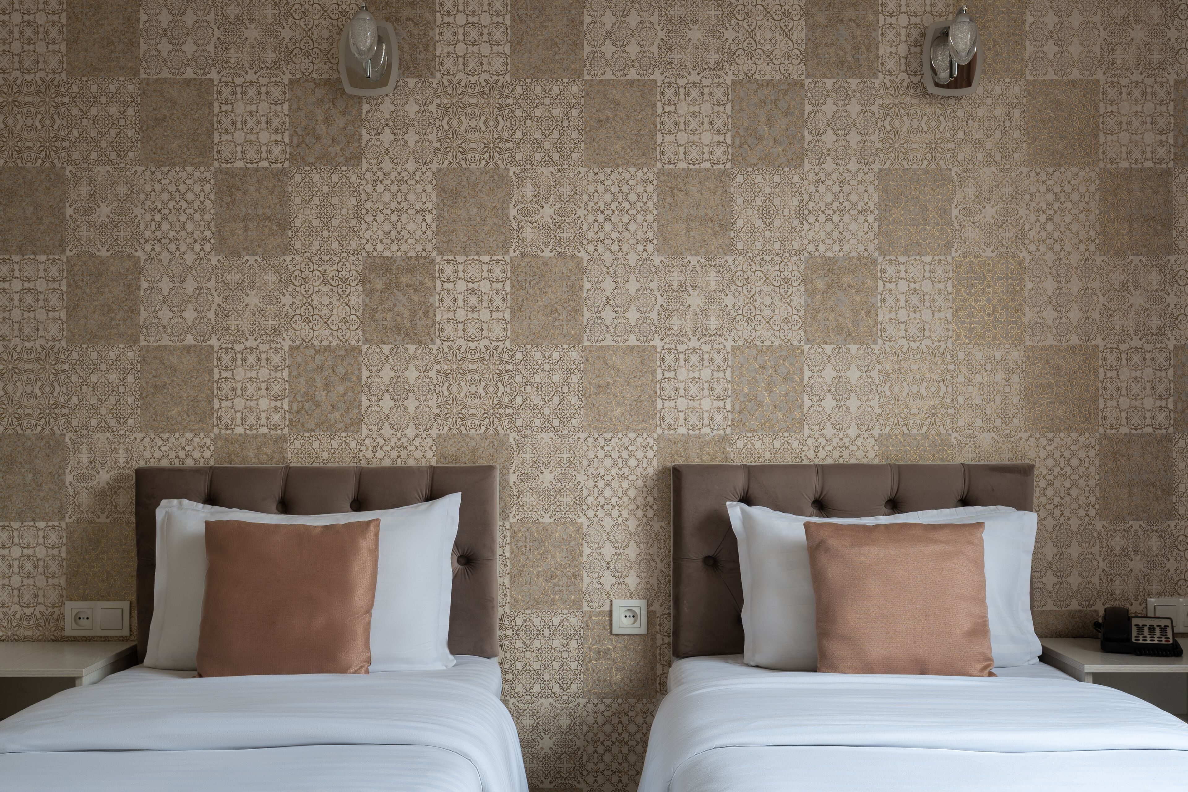 Deux lits jumeaux avec des têtes de lit marron et une literie blanche, un mur décoré d’un motif et une table de chevet avec un téléphone.
