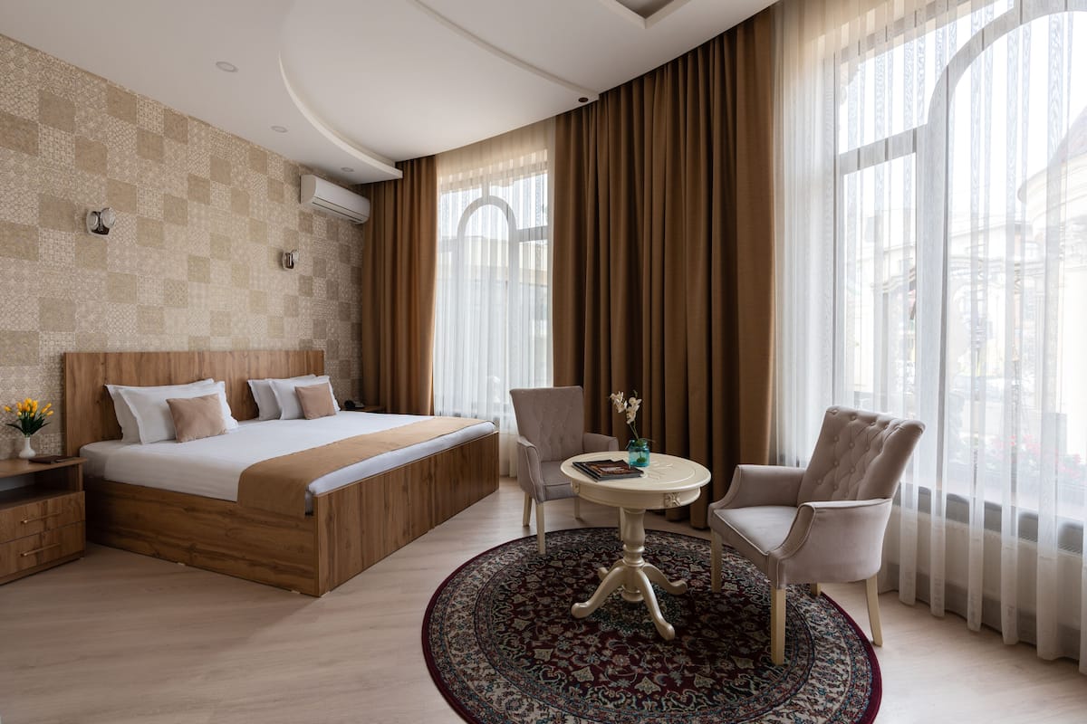 A hotel room with a large bed, two armchairs, a round table, and a view of the city through large windows.