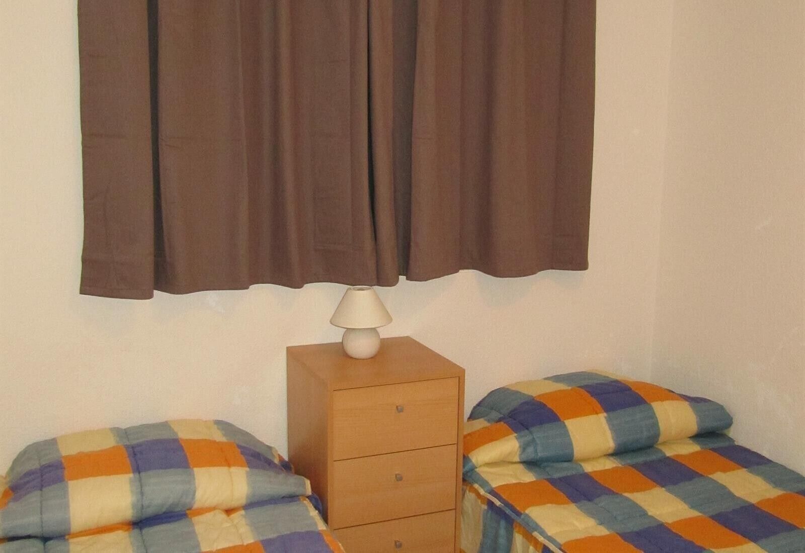 Een kamer met twee enkele bedden, een houten nachtkastje met een lamp en een bruin gordijn.