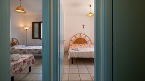 Une chambre avec deux lits, une tête de lit en bois, une croix au mur et une porte donnant sur une autre pièce.