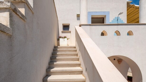 Un bâtiment blanc avec un escalier menant à une porte, une fenêtre et un petit balcon orné d’une plante.