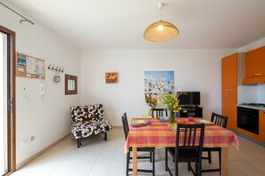 Un coin repas avec une table dressée pour quatre personnes, une chaise à motifs à pois, un luminaire suspendu et un espace cuisine avec des placards orange.