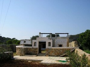 Ein Steinhaus mit einer zentralen Tür, umgeben von einer Steinmauer und einem kleinen Garten mit einem Gartenschlauch.
