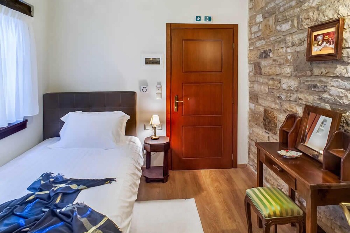 Une chambre d’hôtel avec un lit, une table de chevet avec une lampe, une porte en bois, un mur en pierre et une petite table avec des chaises.