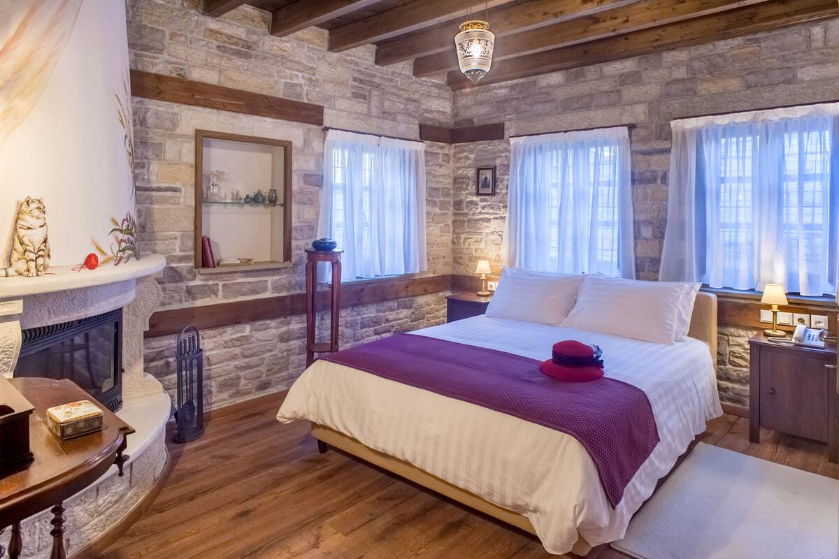 Ein gemütliches Schlafzimmer mit Kamin aus Stein, Holzbalken, einem großen Bett mit rotem Kissen und einem Nachttisch mit Lampe.