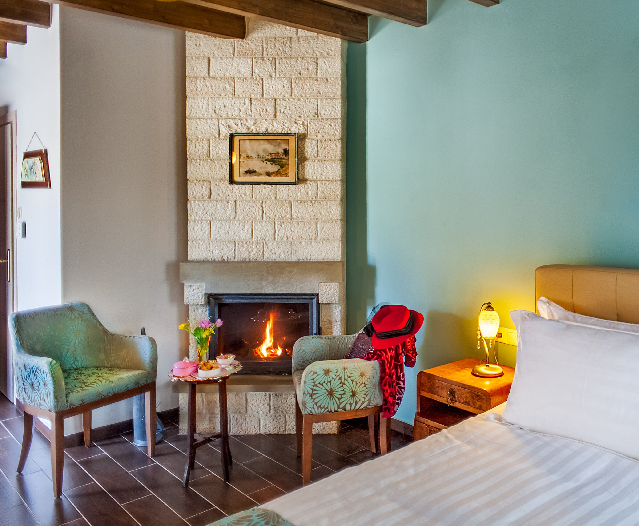 A cozy room with a fireplace, two chairs, a bedside table with a lamp, and a bed.