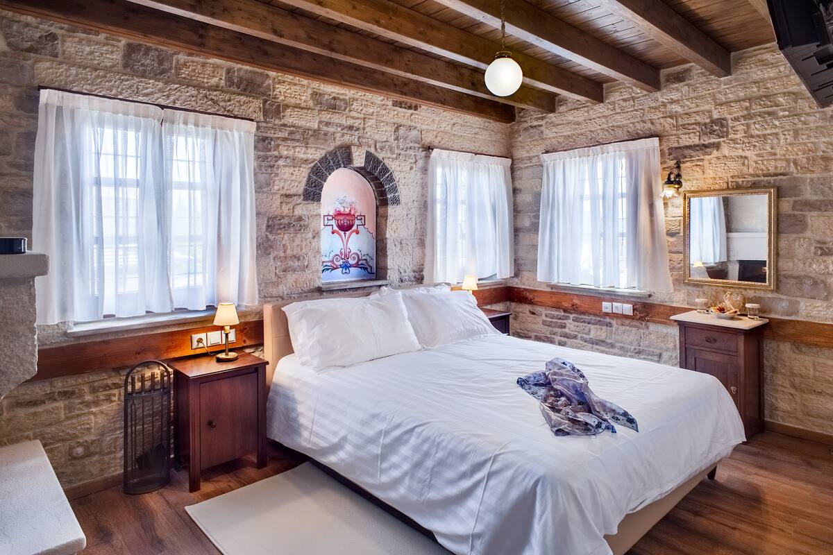 Ein Schlafzimmer mit Steinwand, Holzbalken, einem großen Bett, zwei Nachttischen, einem Schminktisch und einem Fenster mit Vorhängen.
