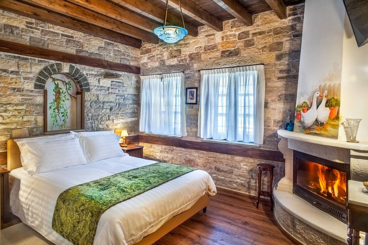 Ein gemütliches Zimmer mit Steinwand, Kamin, einem Bett mit grüner Decke und einem Fenster mit Vorhängen.
