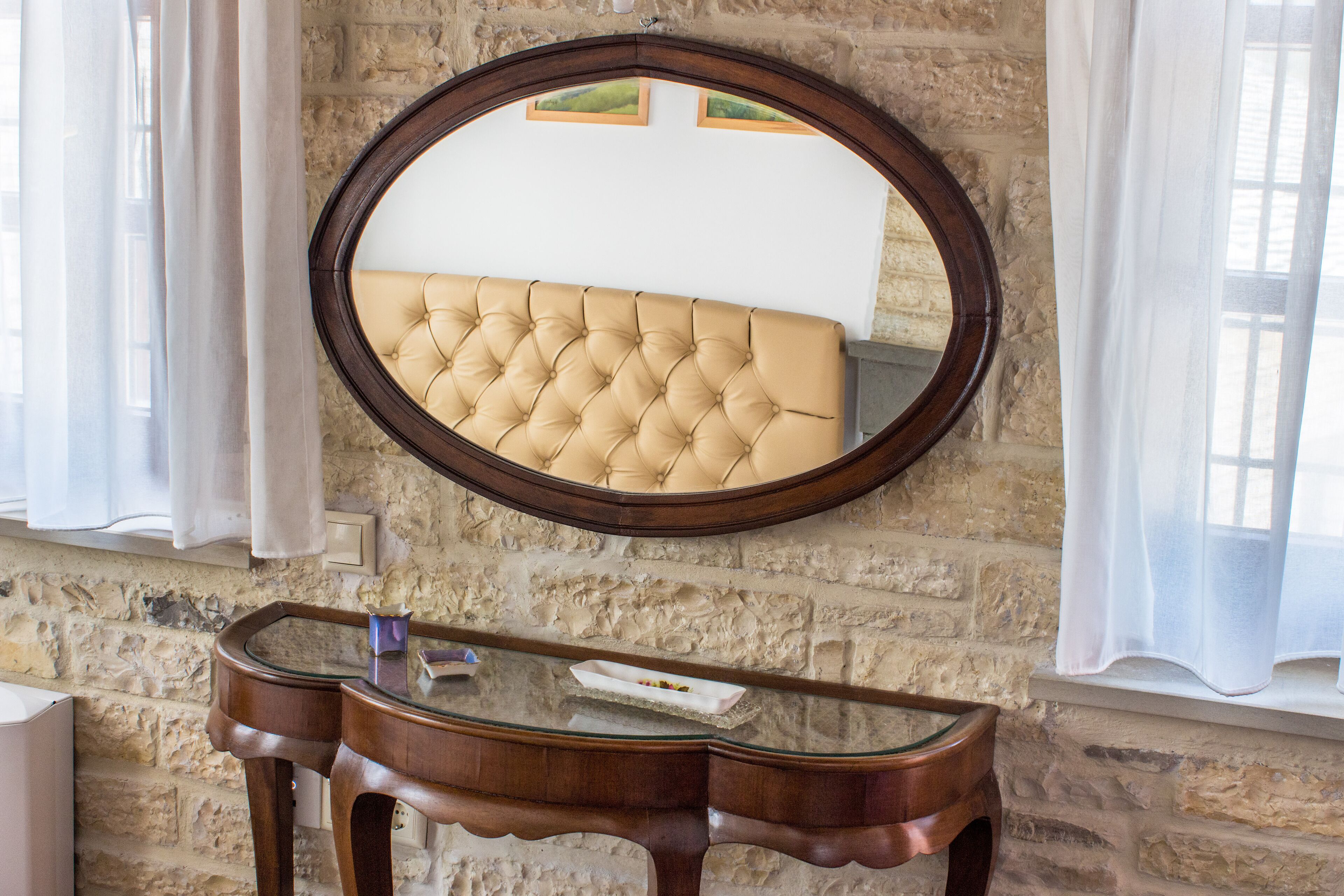 Een kamer met een gestoffeerd hoofdeinde, een ronde spiegel en een houten console met een glazen blad.