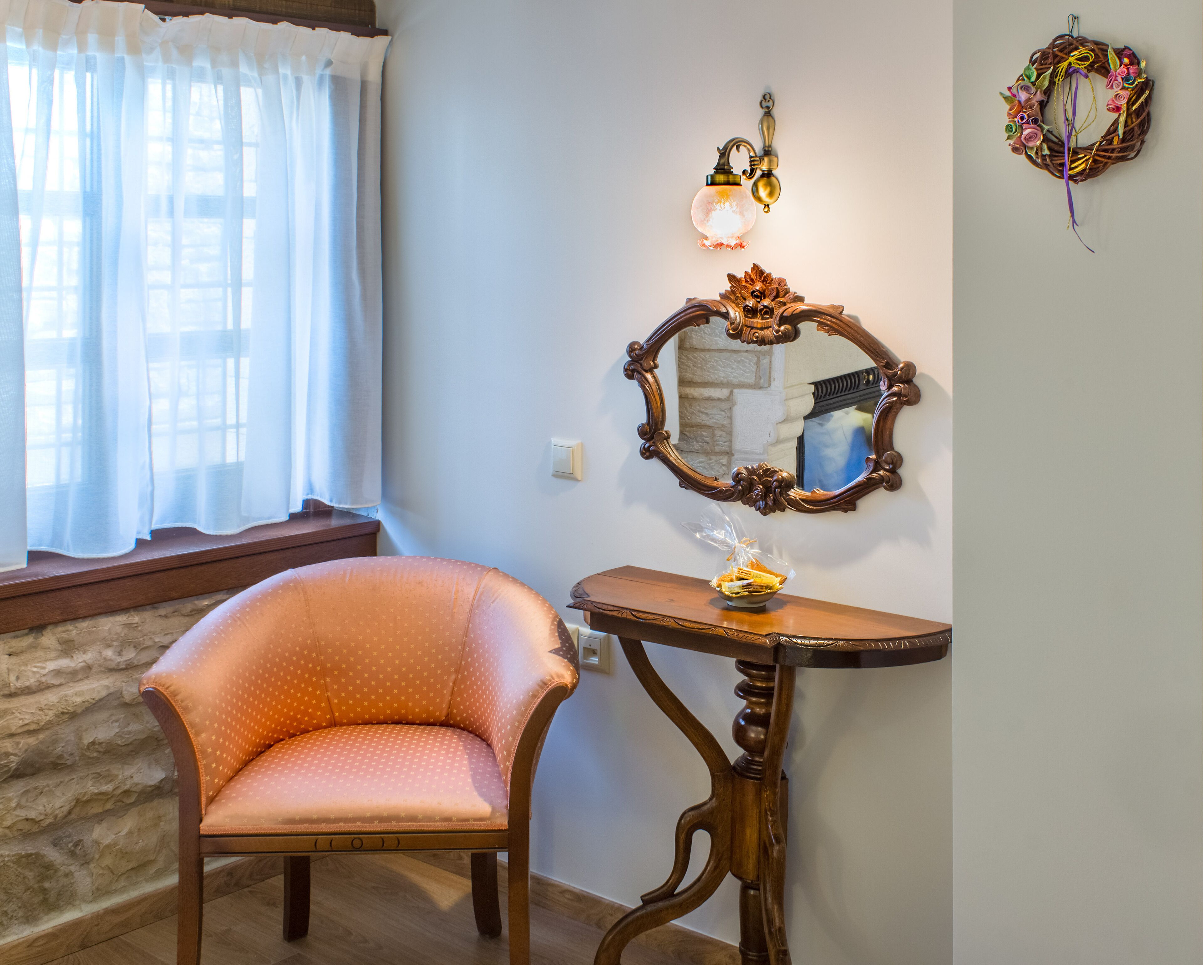 Una habitación con una silla, una mesa con un adorno, un espejo montado en la pared y una ventana con cortinas.