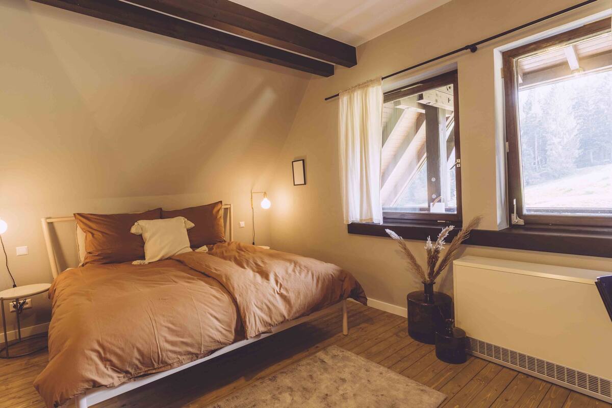 Un dormitorio con cama, mesitas de noche, ventana con cortinas, radiador y suelo de madera.