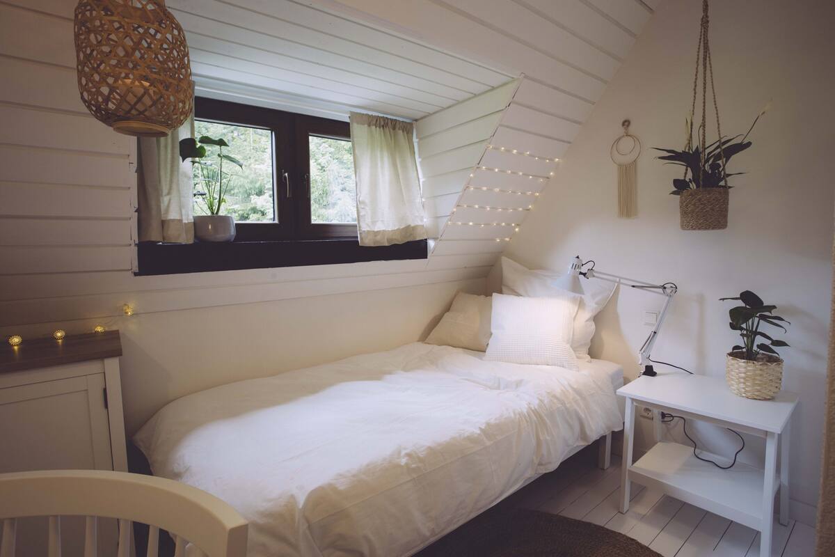 Un dormitorio con cama, mesita de noche, lámpara, plantas colgantes y una ventana con cortinas.
