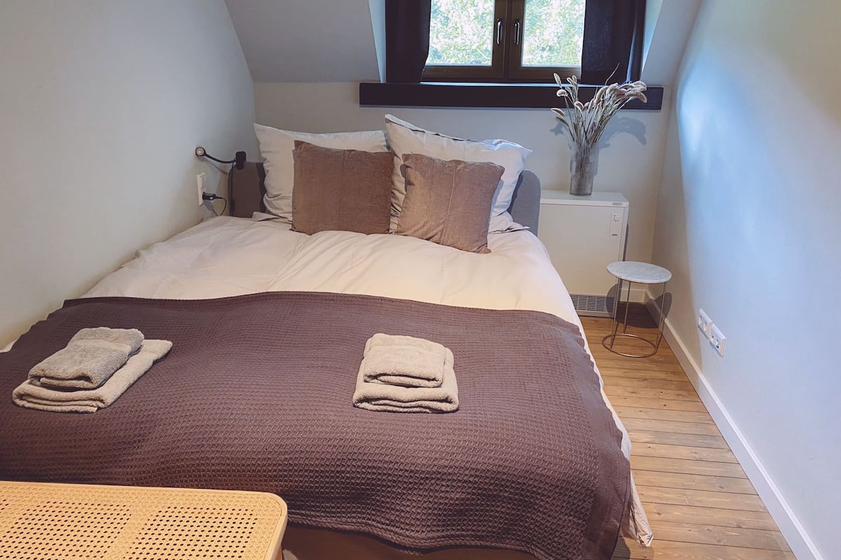 Un dormitorio con cama, almohadas, mesita de noche, radiador y ventana con cortinas.