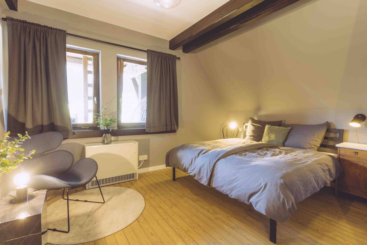 Un dormitorio con cama, una silla, una mesita de noche, una lámpara y una ventana con cortinas.
