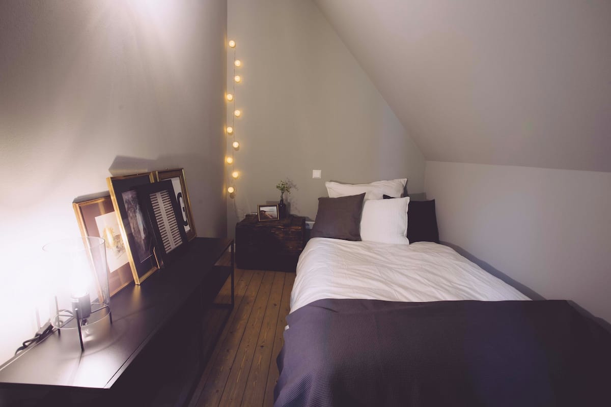 Un dormitorio con techo inclinado, cama con ropa de cama blanca y oscura, mesita de noche con lámpara, y una lámpara de pared.