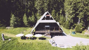 A wooden cabin with a triangular roof, surrounded by a forest. There are outdoor seating areas with umbrellas and a staircase leading to the cabin. - EDELFUCHS-LODGE (Buehlertal)