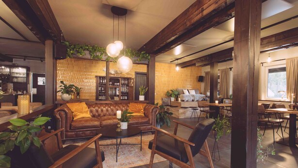 A spacious living area with a leather sofa, wooden chairs, and a coffee table. There are potted plants and a brick wall in the background. - EDELFUCHS-LODGE (Buehlertal)