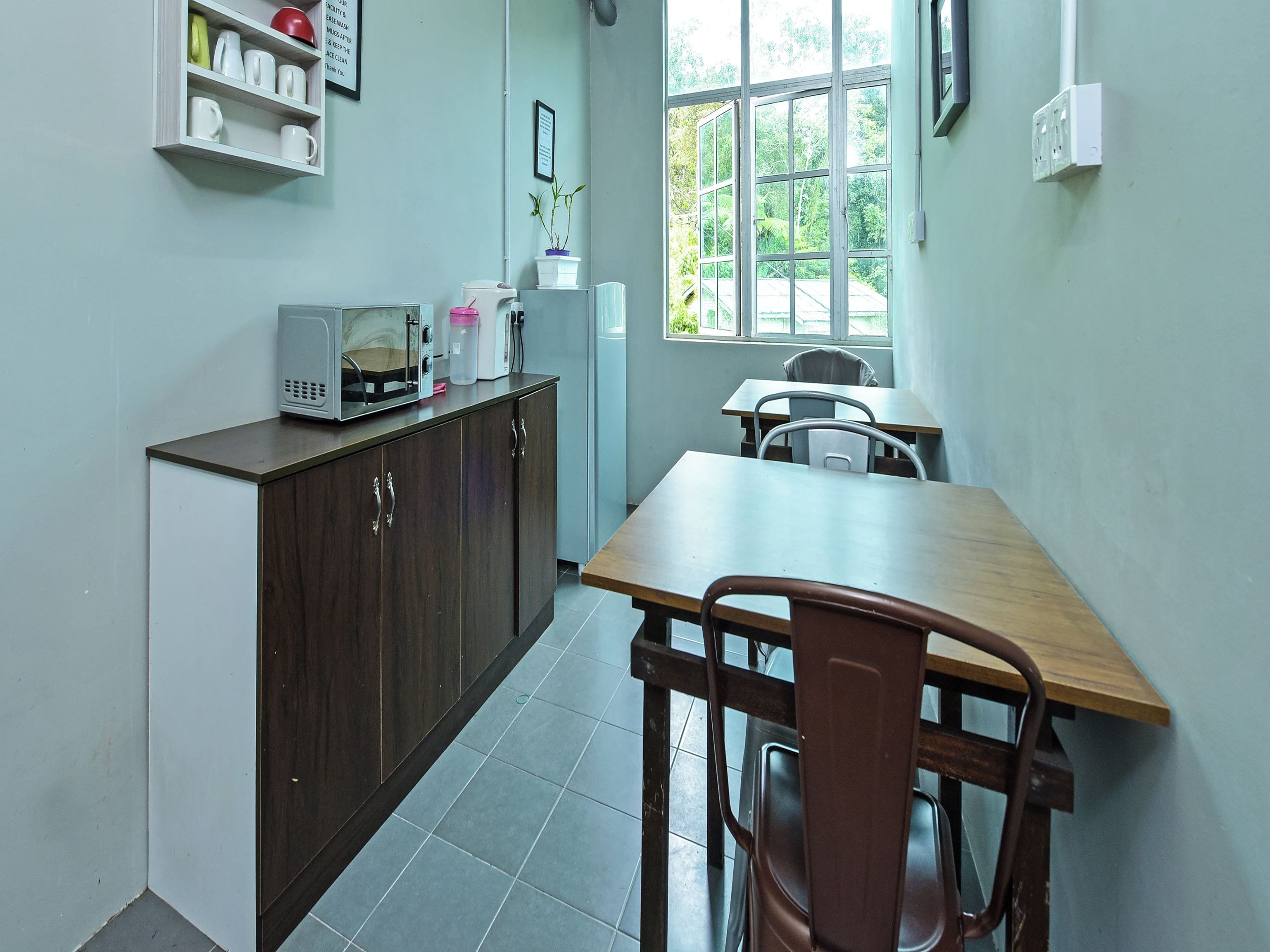 A kitchen with a microwave, refrigerator, and a window with a view of greenery. There is a wooden table and a chair in the dining area.