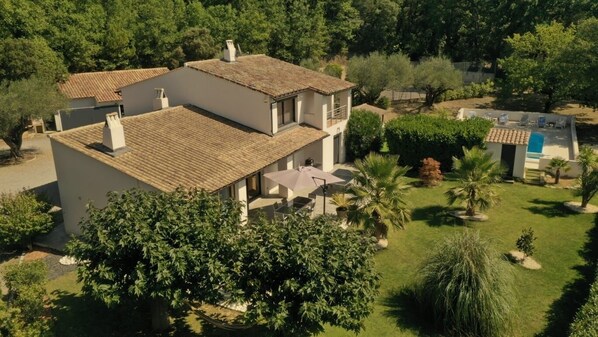 Una casa a due piani con tetto in tegole, circondata da vegetazione lussureggiante e una piccola zona piscina.