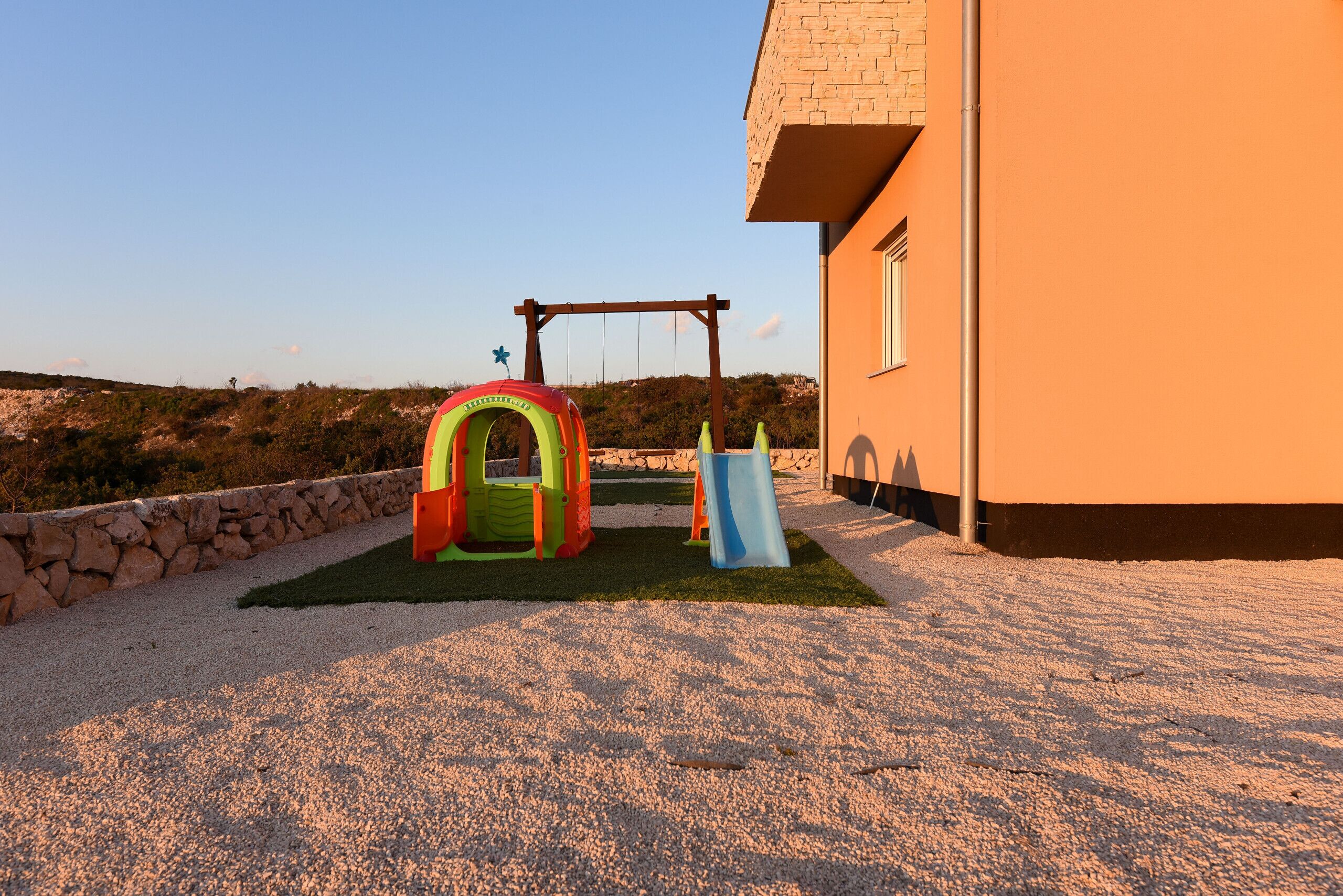 A playground with a slide and a climbing structure, set against a building with a stone wall and a clear sky.