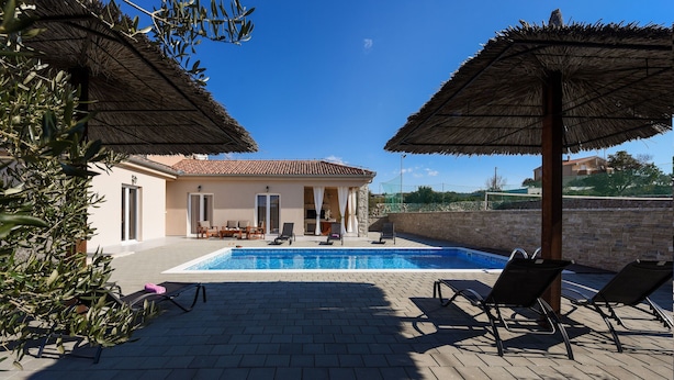 Un espace piscine avec des chaises longues et des parasols, un bâtiment doté d’un toit en tuiles et un ciel d’un bleu limpide.