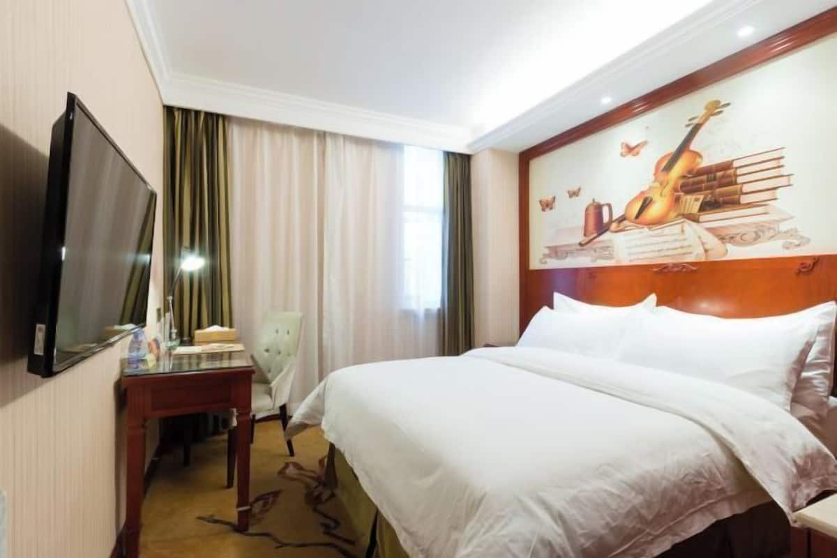 A hotel room with a large bed, a flat-screen TV mounted on the wall, a desk with a chair, and a painting of musical instruments and books on the headboard.