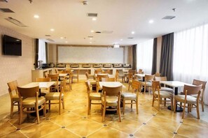 Zona de dinar d'hotel amb diverses taules i caires, una cuina al fons i un gran finestral amb cortines.