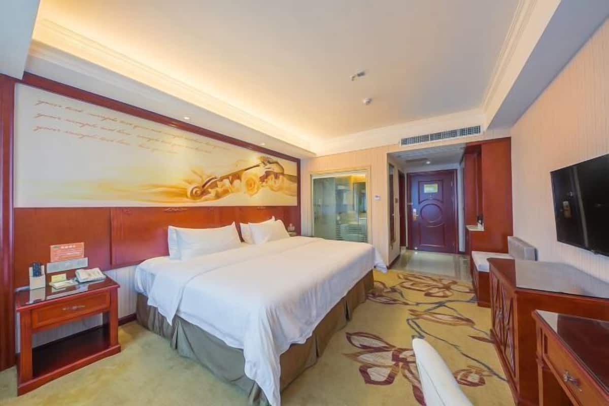 A hotel room with a large bed, a desk with a telephone, a flat-screen TV, and a carpeted floor.