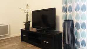 A black entertainment unit with a flat-screen TV, a potted plant, and a tall black lamp. - Modern Apartment for 2-4, 400m from Beach, Terrace, Parking, Quiet Area (Sarzeau)