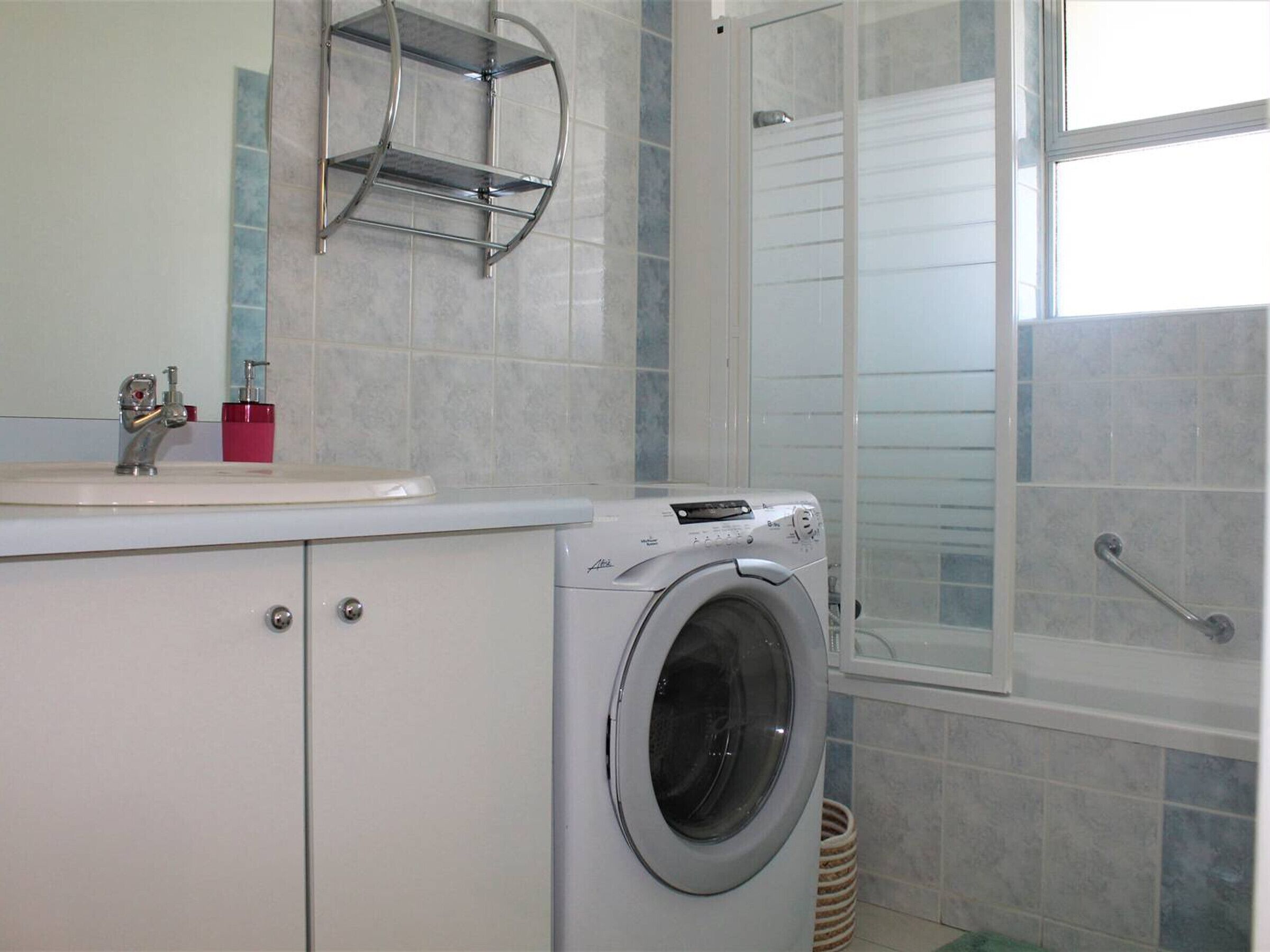 A compact bathroom with a washing machine, a sink with a faucet, a red soap dispenser, and a shower area with a glass door.