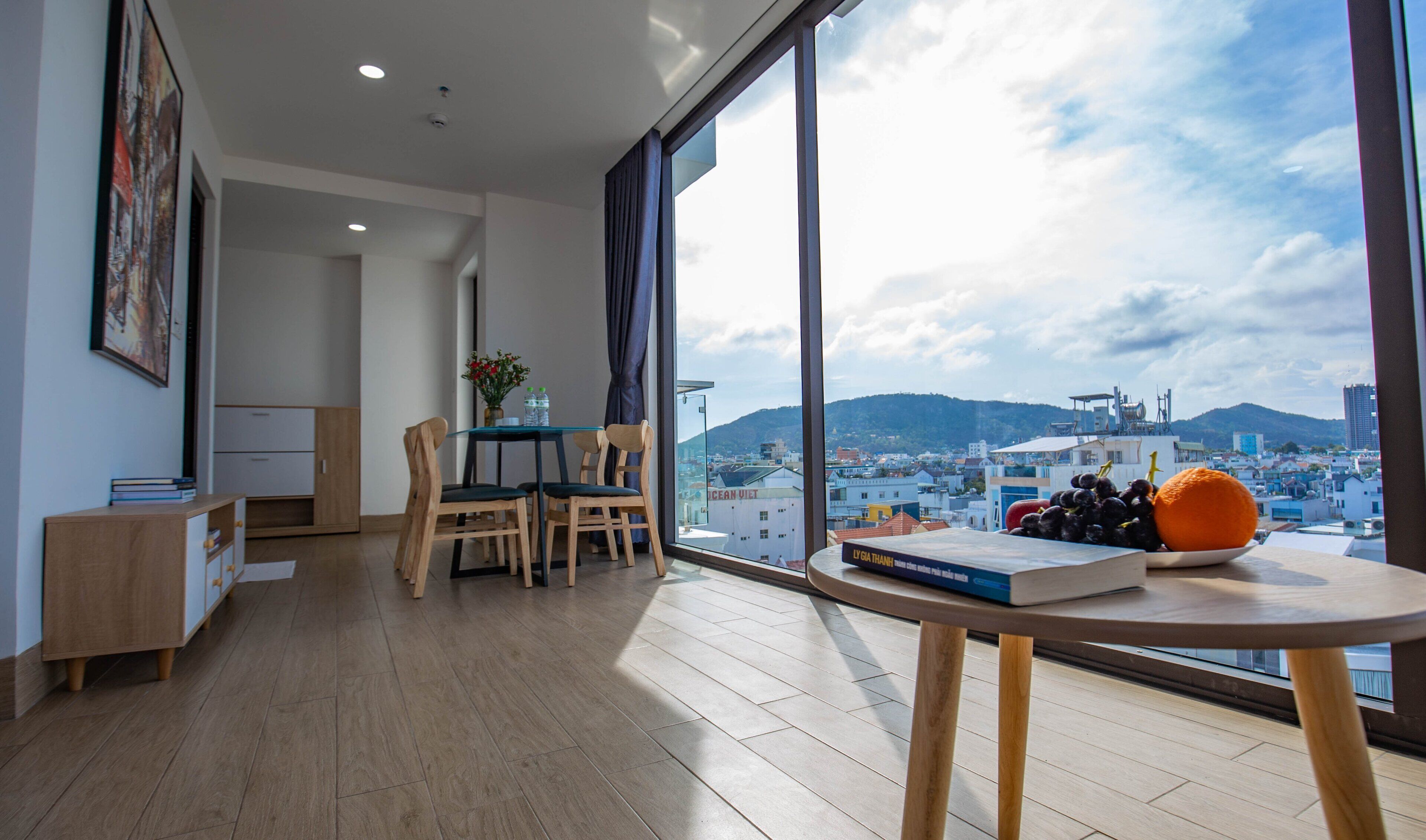 A modern living room with a dining area, large windows with a city view, and a table with fruits and a book.