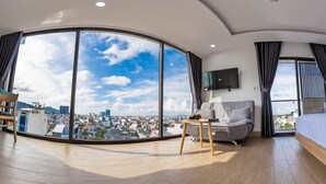 Moderni hotellihuone, jossa on suuri kaareva ikkuna, josta avautuu näkymä kaupunkiin, sekä sohva, sänky ja pieni pöytä, jonka päällä on kaukosäädin.